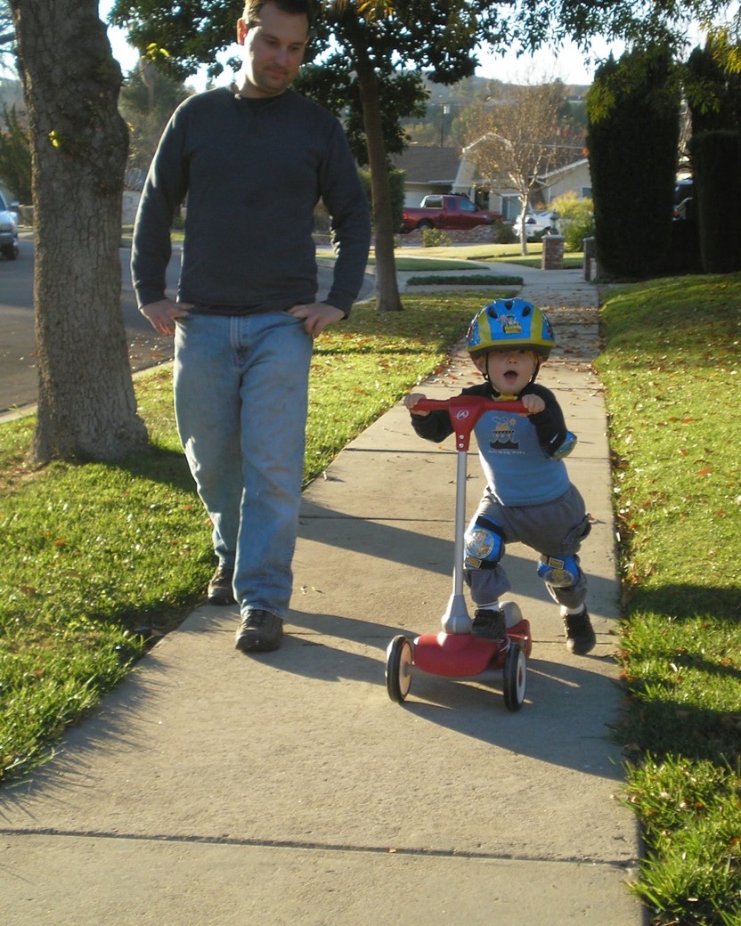 A Bollinger father walking behind his son riding a scooter on their street.