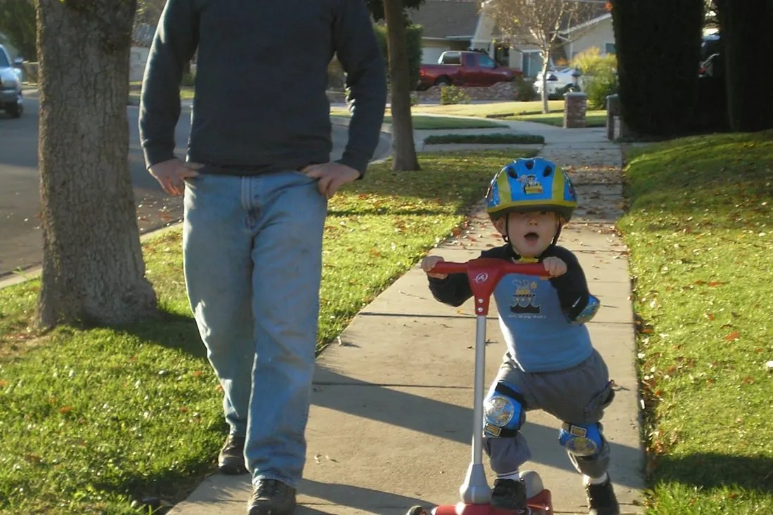 A Bollinger father walking behind his son riding a tricycle.